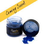 A small clear jar of blue pigment powder with a black lid placed beside it, with a spilled amount of vivid blue powder in front. A yellow diagonal banner across the top reads 'Coming Soon!' in black script.